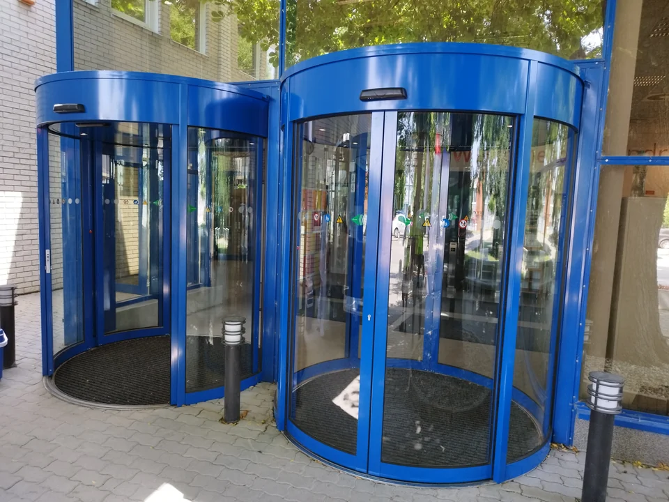 Két eltérő átmérőjű, kék keretes forgóajtó egymás mellett egy épület bejáratánál